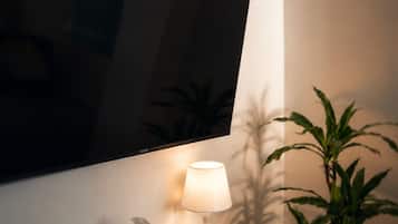Een flatscreen televisie aan een muur, een nachtkastje met een lamp, een afstandsbediening, een zakdoekendoos en een potplant.