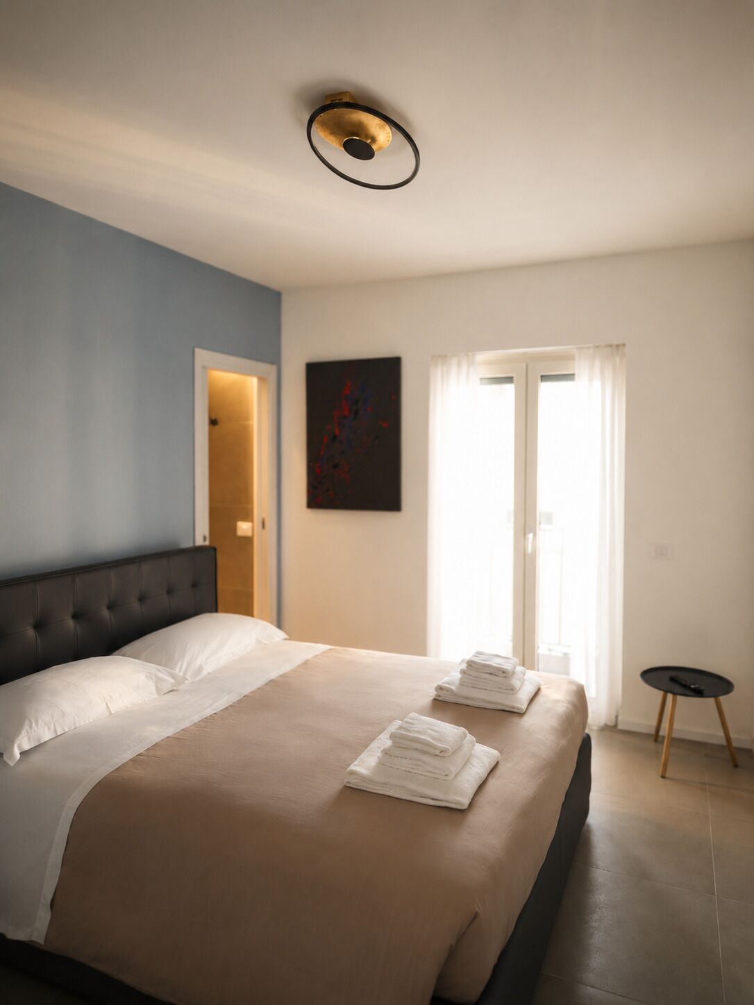 A hotel room with a large bed, a headboard, a painting on the wall, a door, and a small table.