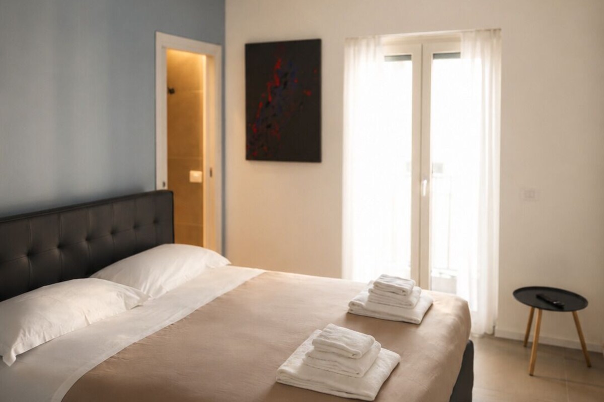 Una habitación de hotel con una cama grande, un cabecero, un cuadro en la pared, una puerta y una mesita.