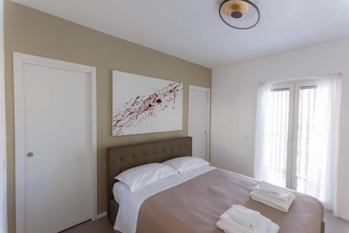 A hotel room with a bed, a large abstract painting, a window with curtains, and a door.