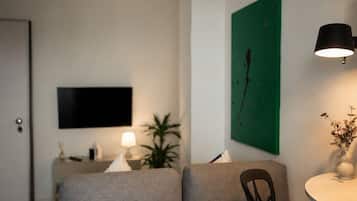 A modern living room with a sofa, a flat-screen TV mounted on the wall, a small table with a lamp, and a green abstract painting.