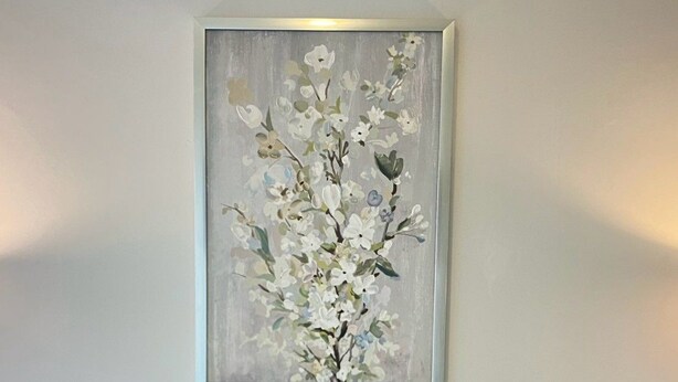 Un sofá blanco con dos almohadas azules y una anaranjada. Encima del sofá, hay una obra de arte floral enmarcada en la pared.