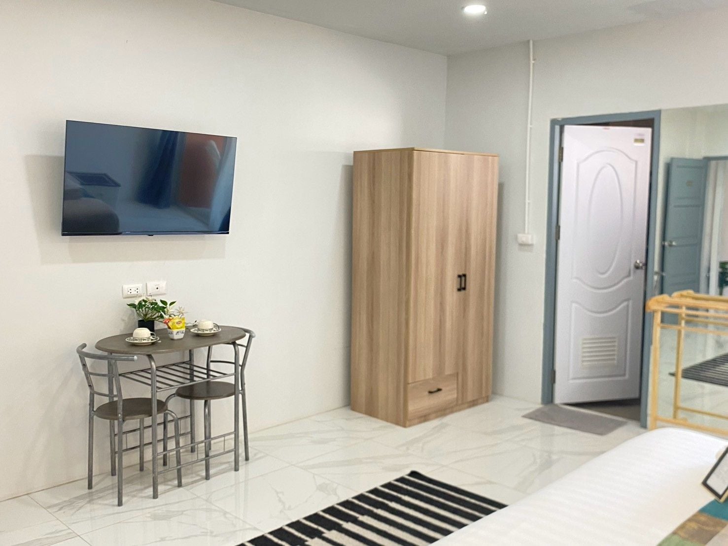 Une chambre moderne dotée d’un téléviseur à écran plat fixé au mur, d’une armoire en bois, d’une petite table ronde avec des chaises et d’une porte avec une poignée.
