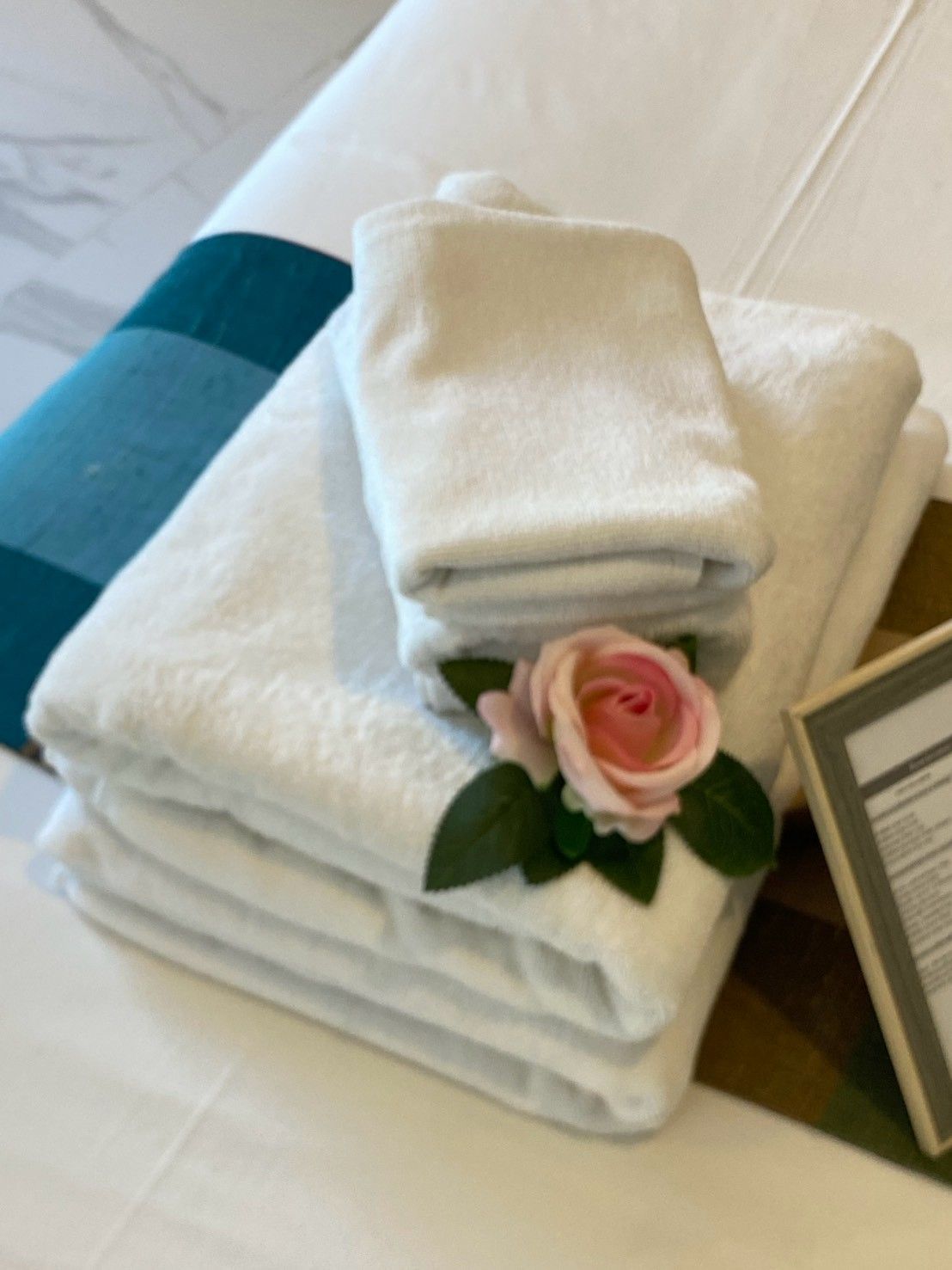 A stack of white towels with a pink rose on top, placed on a bed with a white pillow and a framed picture beside them.