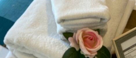 A stack of white towels with a pink rose on top, placed on a bed with a white pillow and a framed picture beside them.