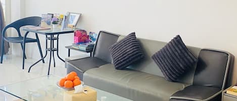 A modern living room with a glass coffee table, a grey sofa with black cushions, a wall art piece, and a dining area with a chair and table.