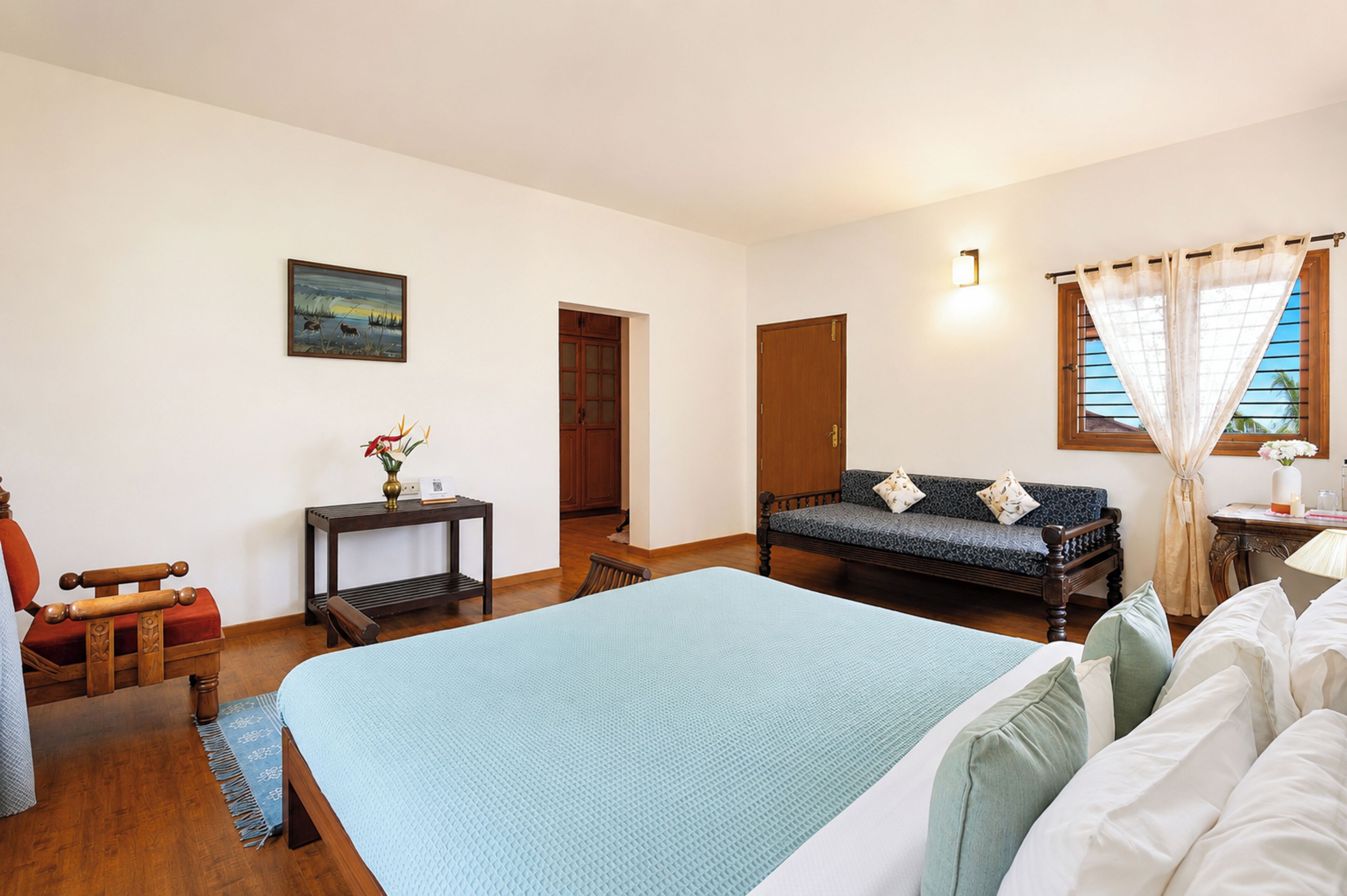 A room with a bed, a sofa, a chair, a small table with a vase, and a painting on the wall.