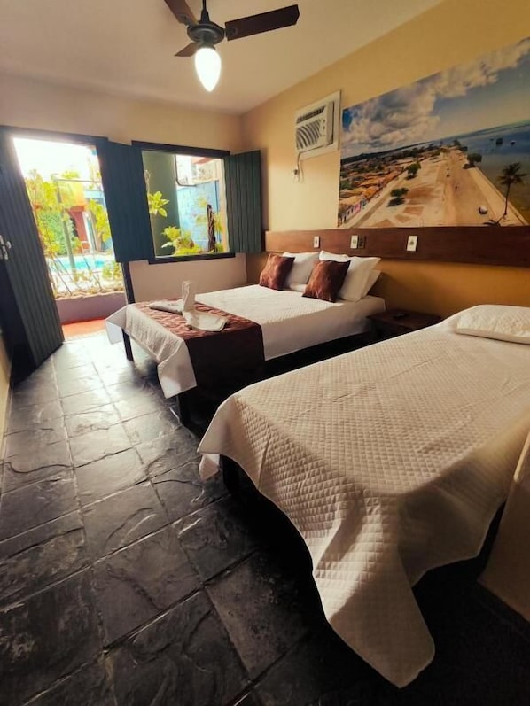 A hotel room with two beds, a ceiling fan, a painting of a beach, and a view of the outdoors.