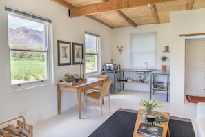 A spacious room with a wooden dining table, chairs, a kitchen area, and large windows offering a view of a green landscape.