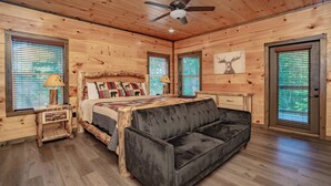 A wooden cabin room with a bed, a black sofa, a ceiling fan, and wooden walls. - SmokySplashRetreat: Pool, Wifi, Hot Tub, Game Room, Partial Mtn Views (Sevierville)