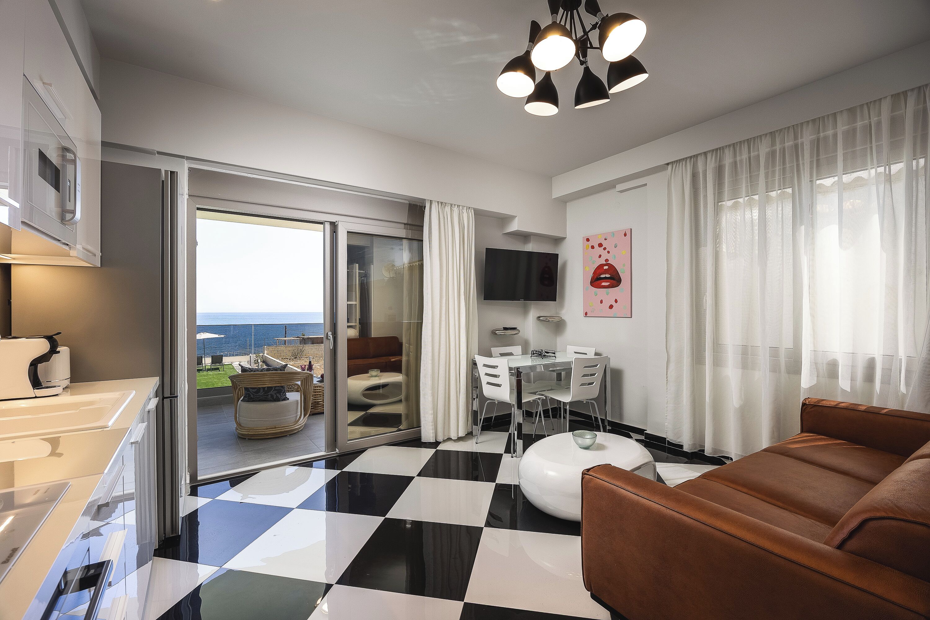 A modern living room with a checkered floor, a brown sofa, a dining table with chairs, a TV mounted on the wall, and a sliding glass door leading to a balcony.