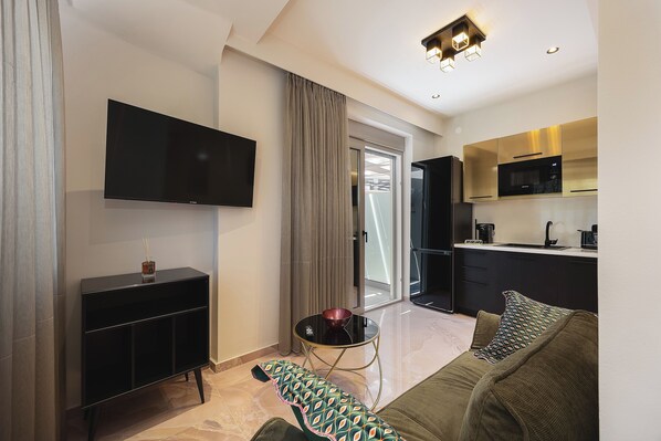 A modern living room with a flat-screen TV, a sofa, a coffee table, and a kitchen area with a microwave. - MGM Suites (Mylopotamos)