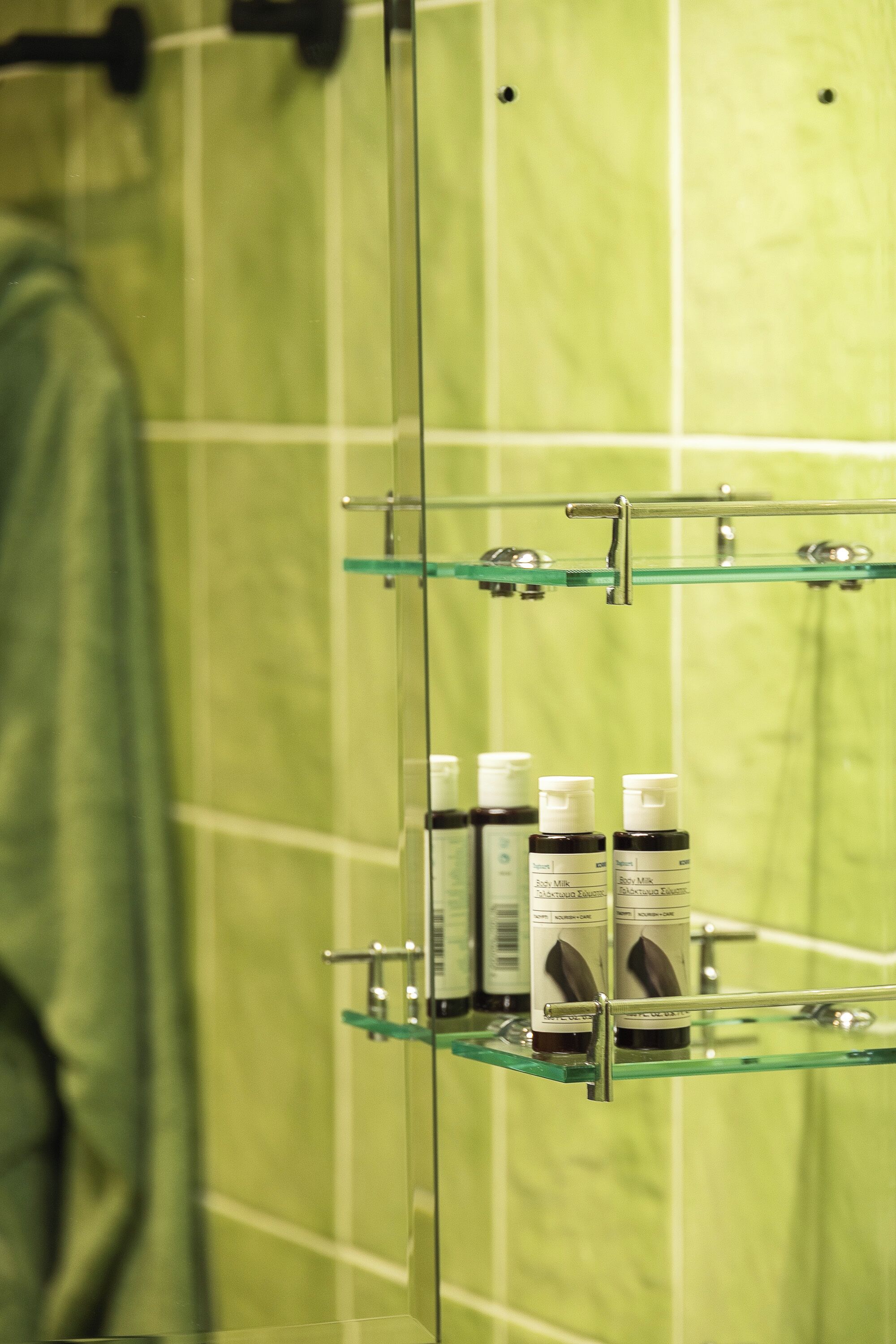 Een badkamer met groene tegels, een glazen douchecabine en twee glazen planken met flessen.
