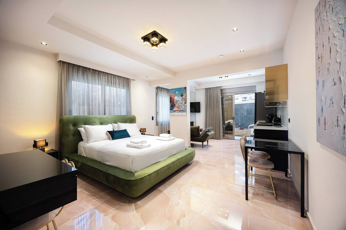 A modern hotel room with a green upholstered bed, a desk with a chair, a flat-screen TV, and a dining area.