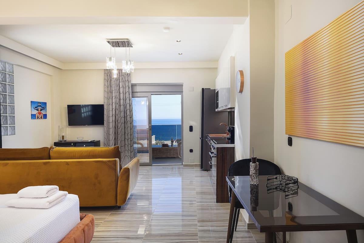 A hotel room with a sofa, a TV, a dining table, and a view of the ocean.