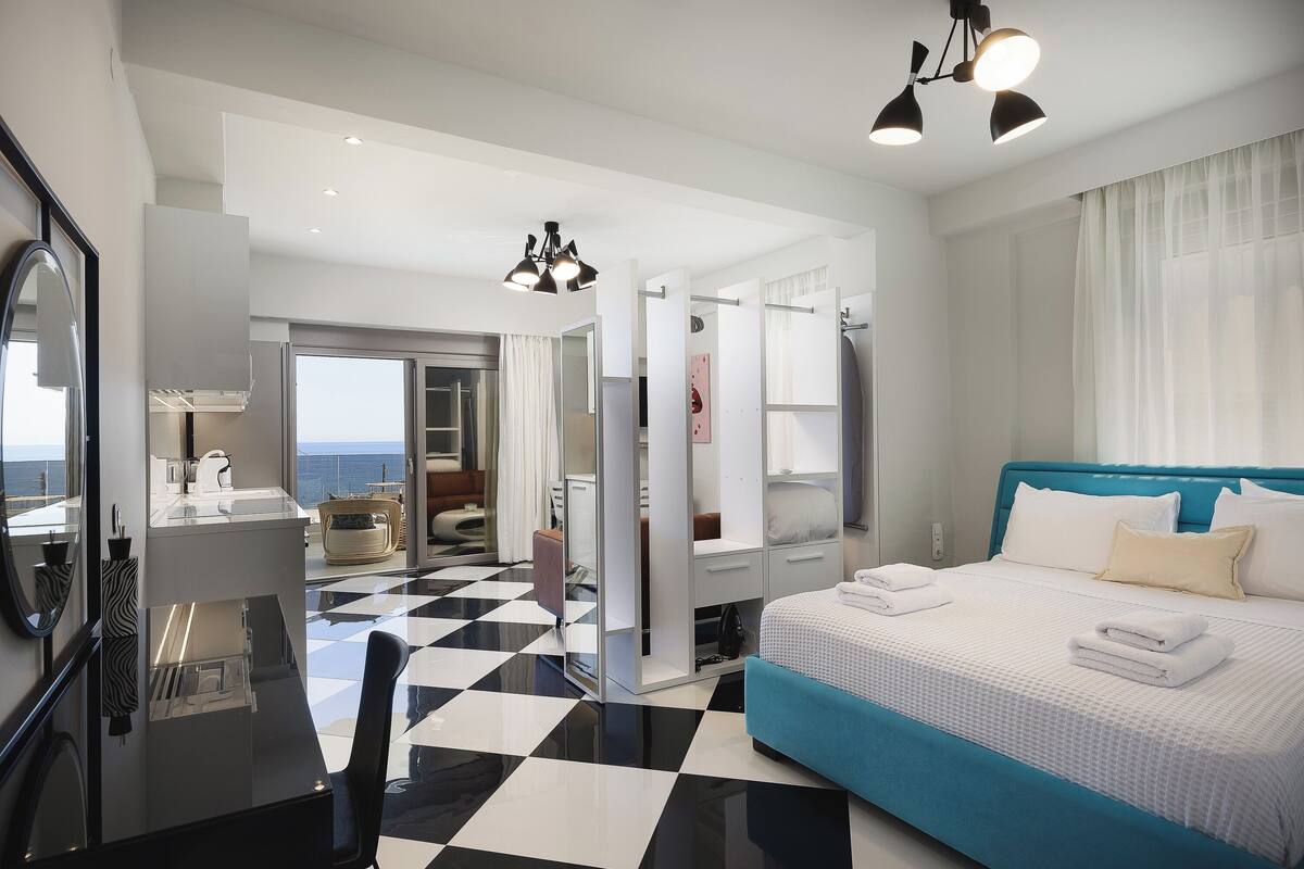 A hotel room with a checkered floor, a bed with blue headboard, a desk with a mirror, and a view of the sea.