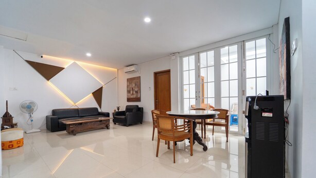 A modern living room with a sofa, a coffee table, a dining table with chairs, a TV, and large windows. - Tempuran Suites By ARBA (Sleman)