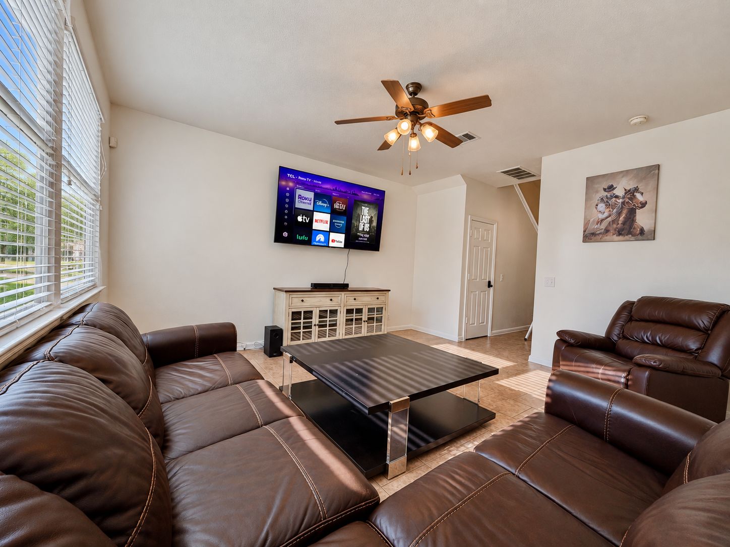 A living room with a brown leather sofa, a coffee table, a ceiling fan, and a flat-screen TV mounted on the wall.