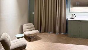 Ein Zimmer mit Fischgratparkett, einem gepolsterten Sessel, einem kleinen runden Tisch, einem Fenster mit Vorhang und einer Küchenzeile.
