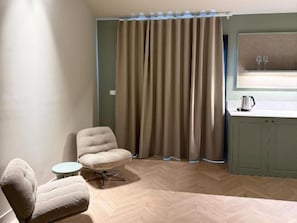 Ein Zimmer mit Fischgratparkett, einem gepolsterten Sessel, einem kleinen runden Tisch, einem Fenster mit Vorhang und einer Küchenzeile.