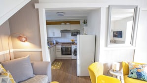 Een knus woongedeelte met een kitchenette, een bank, een gele fauteuil en een tafeltje met een lamp.