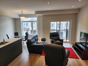 A modern living room with a sofa, armchair, TV, and a glass table. - Modern, bright and private 2 bedroom, 2 full  bathroom (Cambridge)