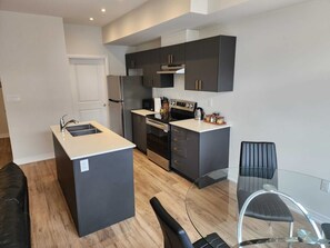 A modern kitchen with dark cabinetry, stainless steel appliances, a glass dining table, and a black leather sofa. - Modern, bright and private 2 bedroom, 2 full  bathroom (Cambridge)