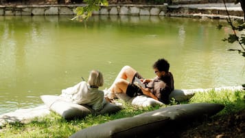 Dos personas sentadas en cojines junto a un estanque, con césped y árboles al fondo.