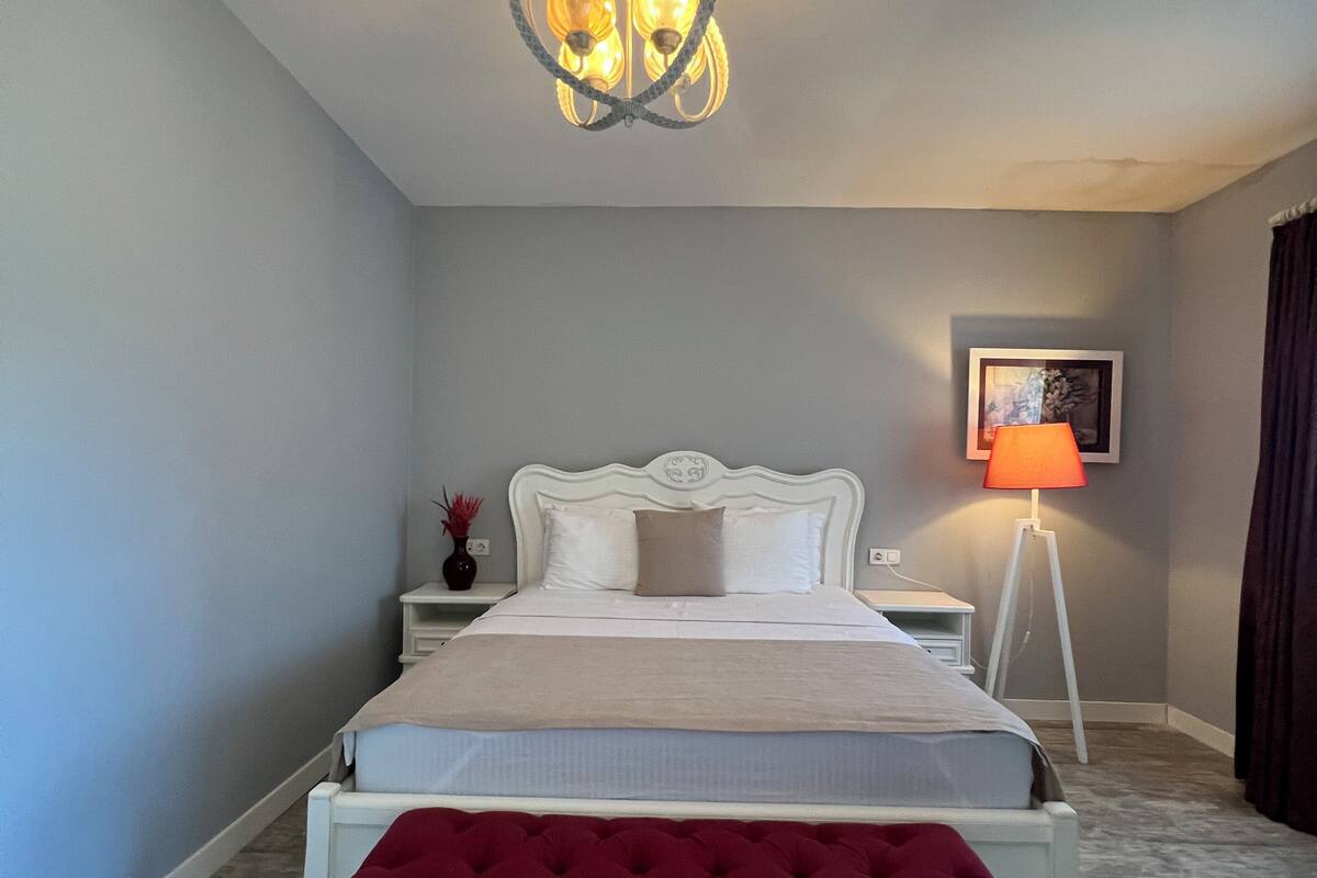 A bedroom with a bed, a red bench, a nightstand, a floor lamp, and a chandelier.