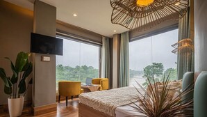 A modern hotel room with a large bed, a desk with a laptop, a TV, and a view of the outdoors. - River View Resort (Haridwar)