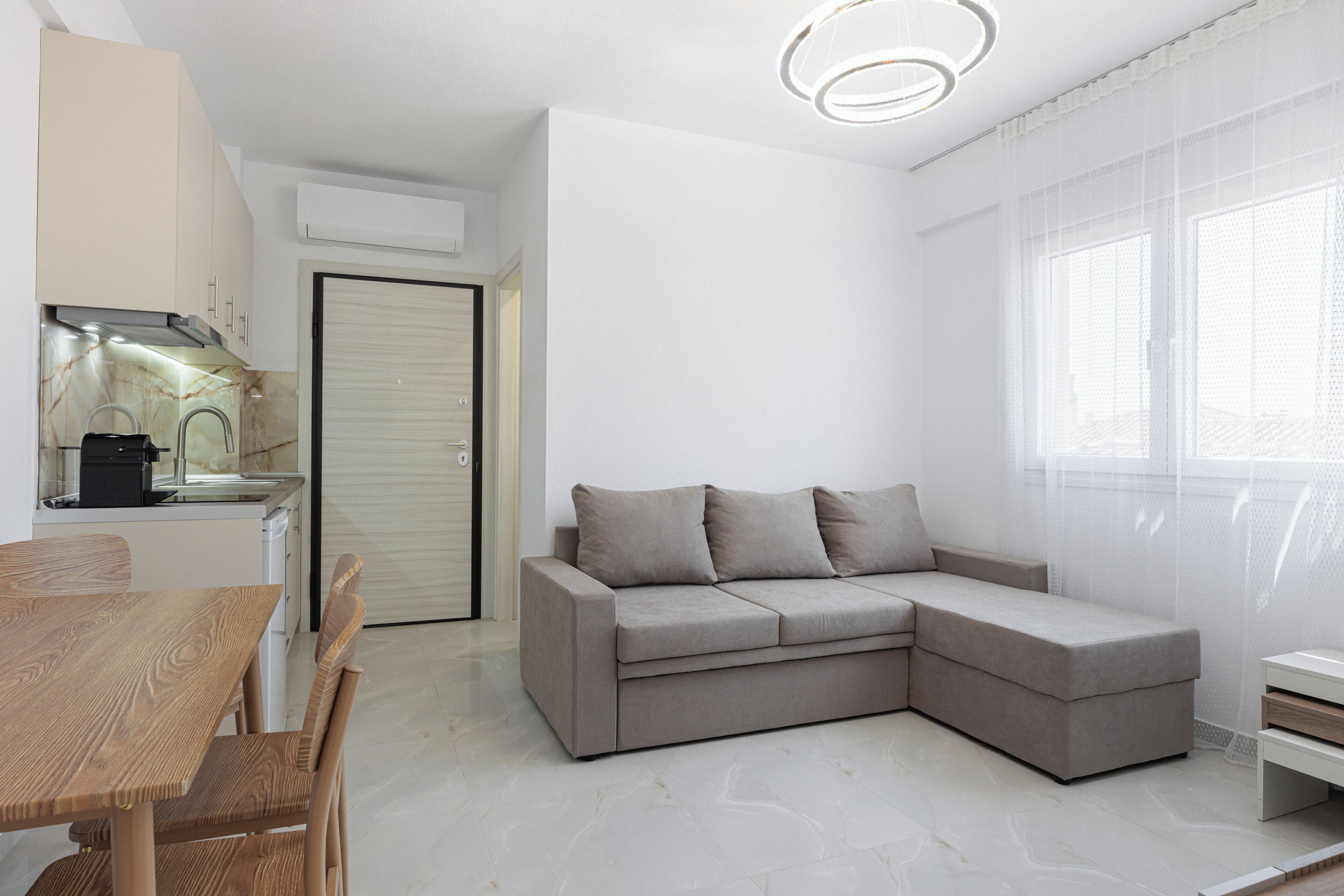 A modern kitchen with a built-in stove, a dining table with chairs, a grey sectional sofa, and a large window with sheer curtains.
