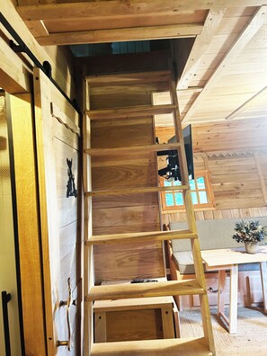 Interior de madera con una escalera que lleva a un altillo, una puerta de madera con picaporte y una ventana con vidrio azulado.