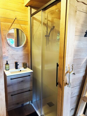Interior em madeira com chuveiro em cabine de vidro, espelho redondo e pia com produtos de higiene.