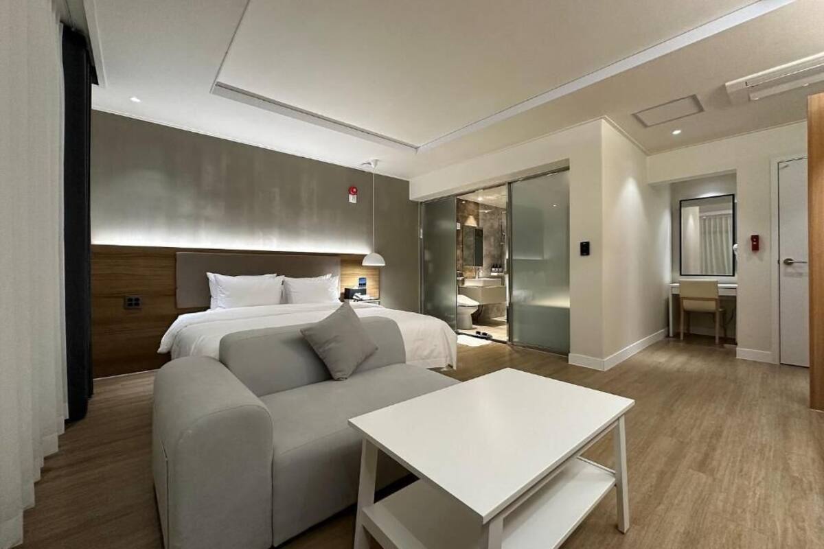 A modern hotel room with a bed, a sofa, a table, and a bathroom visible through a glass door.