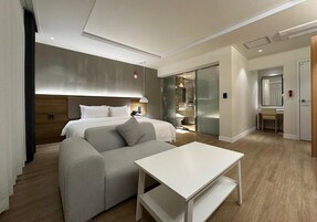 A modern hotel room with a bed, a sofa, a table, and a bathroom visible through a glass door.