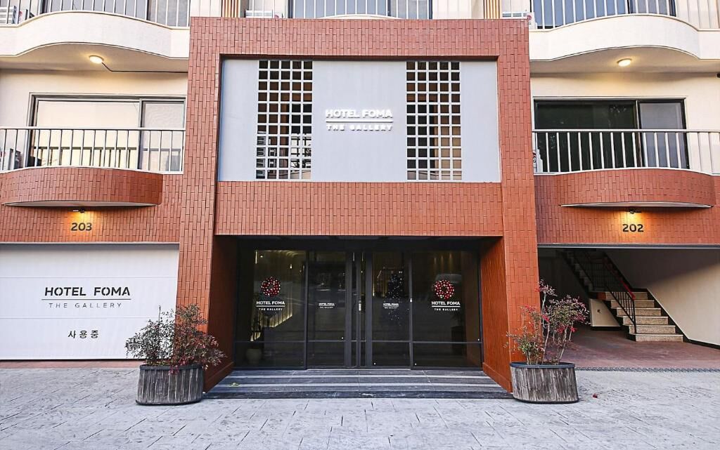 A hotel entrance with a modern design, featuring a glass facade and a sign that reads 'HOTEL FOMA THE GALLERY'.