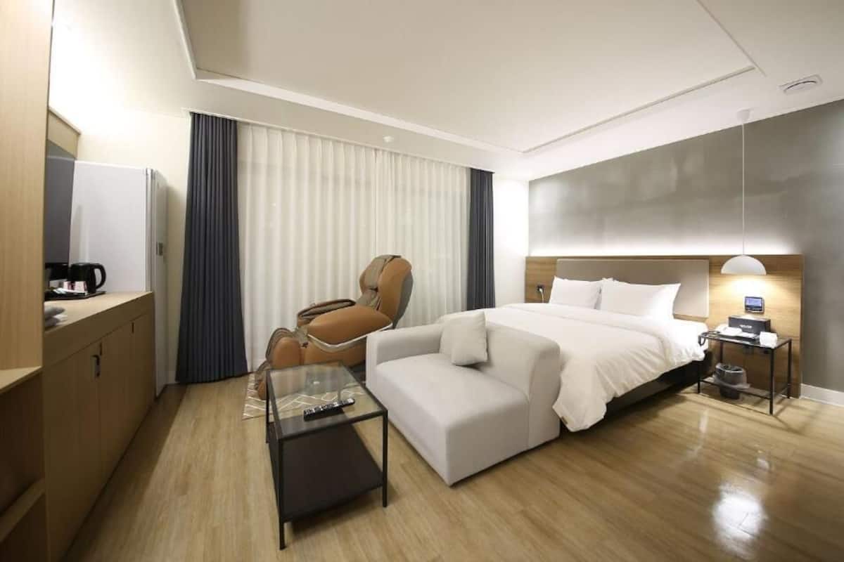 A modern hotel room with a bed, a sofa, a chair, a coffee table, and a desk with a coffee maker.