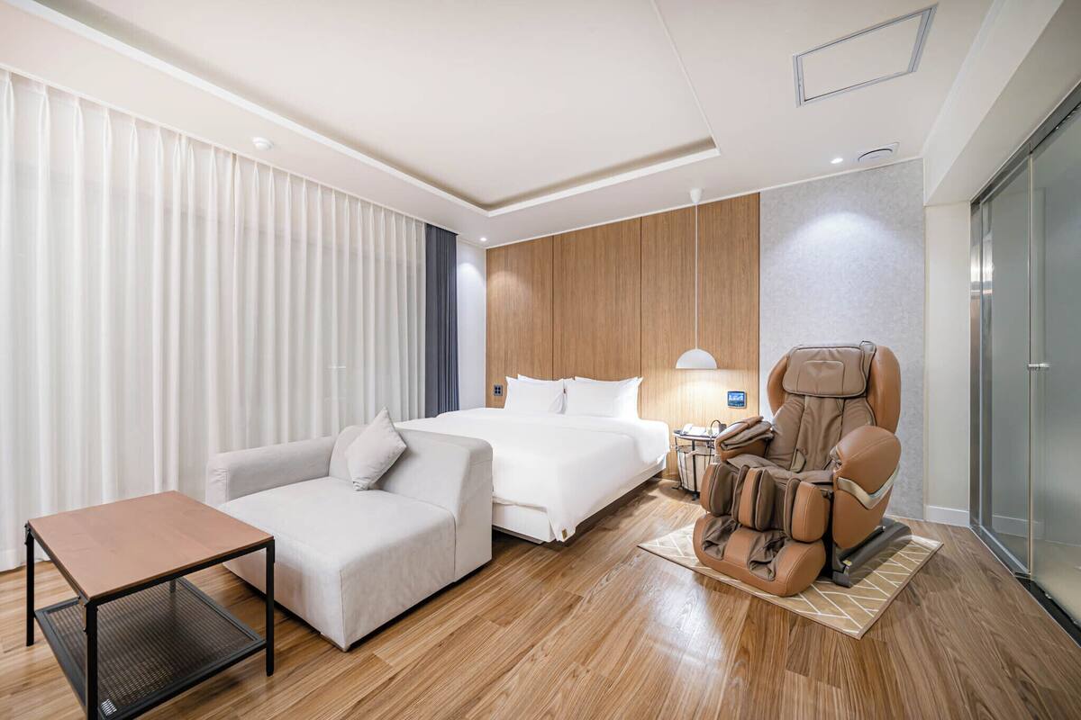 A hotel room with a bed, a sofa, a massage chair, a small table, and a glass partition.
