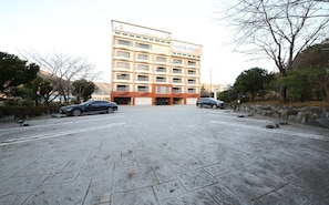 A multi-story hotel with a flat facade, a parking lot with two cars, and a stone-paved entrance. - Hotel Foma (Changwon)