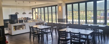 A restaurant with tables and chairs, a counter with appliances, and large windows with a view.