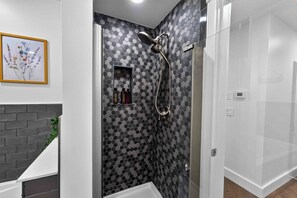 A modern bathroom with a shower, a framed floral artwork, and a shelf with toiletries. - Heron's Rest – Lakefront Home w/ Hot Tub, Kayaks & Game Room (Pocono Lake)