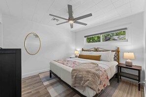 Quarto com cama, ventilador no teto, janela com vista, mesa de cabeceira com abajur e espelho na parede.