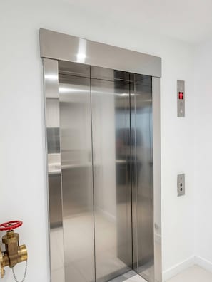 Een moderne lift met een metalen afwerking, een brandmeldknop en een bedieningspaneel.