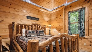 Un dormitori de cabana de fusta amb un llit, una tauleta de nit, una làmpada i una finestra amb cortines.