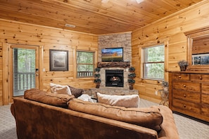 Ein Holzblockhaus-Innenraum mit Kamin aus Stein, einem darüber montierten Fernseher und einem braunen Ledersofa.