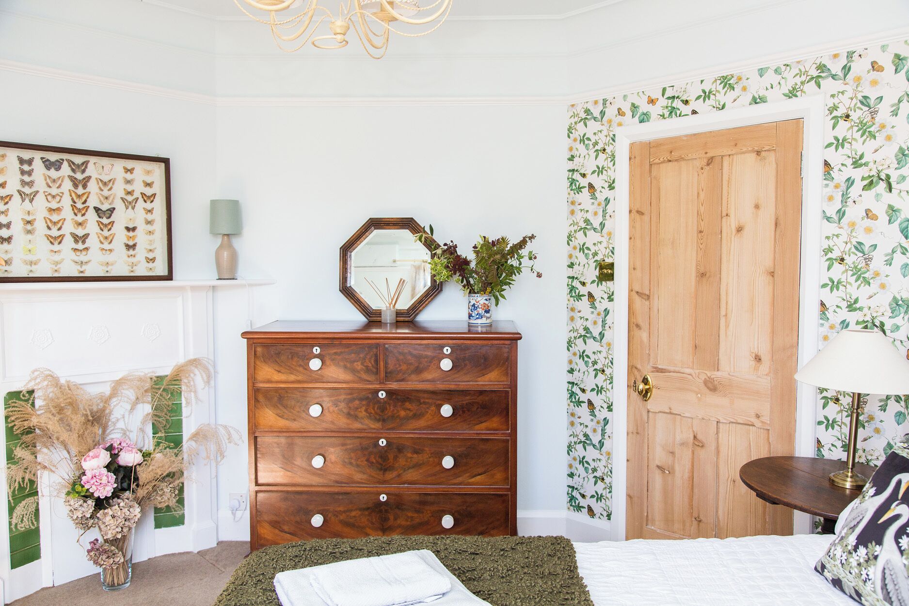 A bedroom with a wooden dresser, a mirror, a floral wallpaper, a door, and a bed with a green blanket.