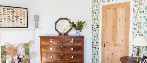 A bedroom with a wooden dresser, a mirror, a floral wallpaper, a door, and a bed with a green blanket.