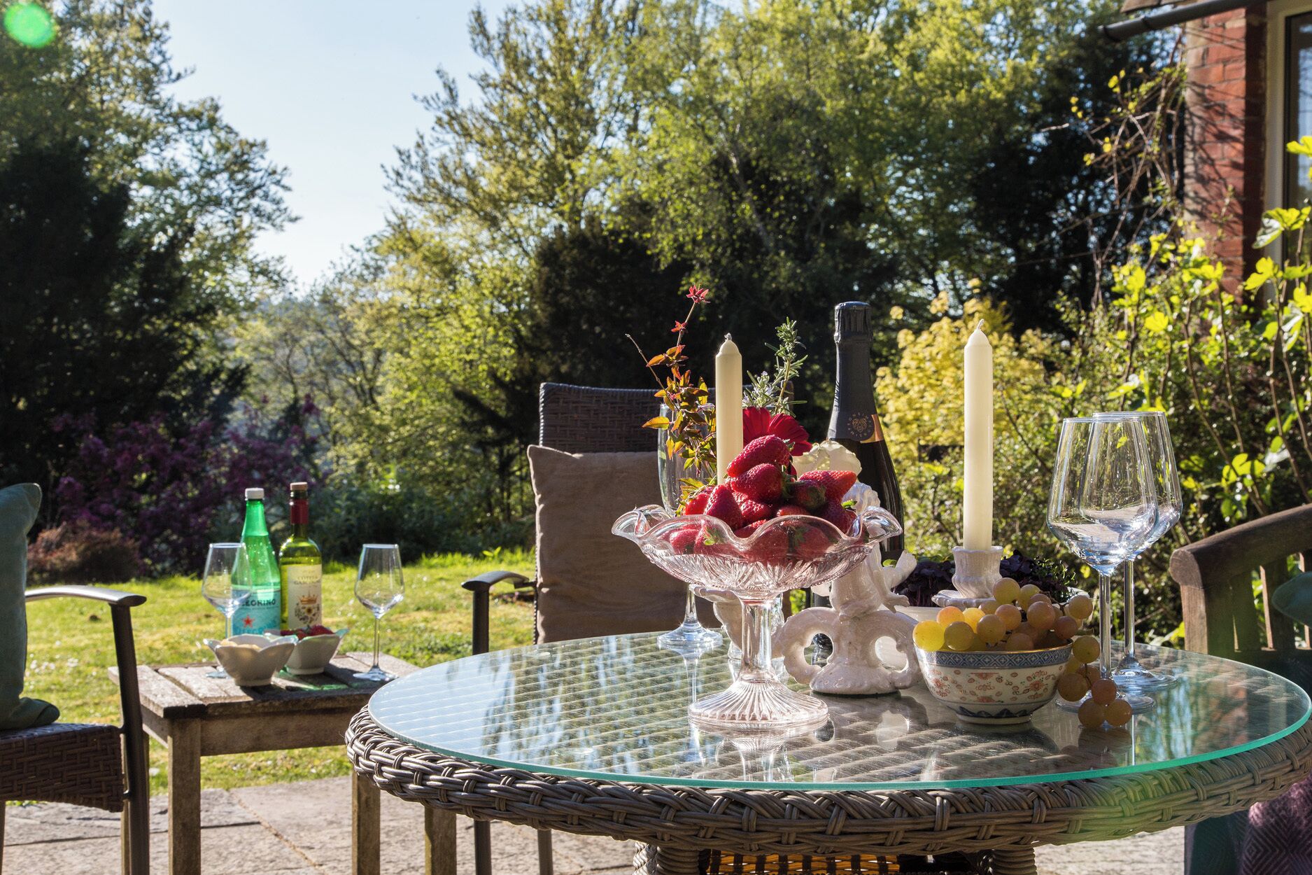 A round glass table with wicker chairs, a bowl of strawberries, a candle, and a bottle of wine.