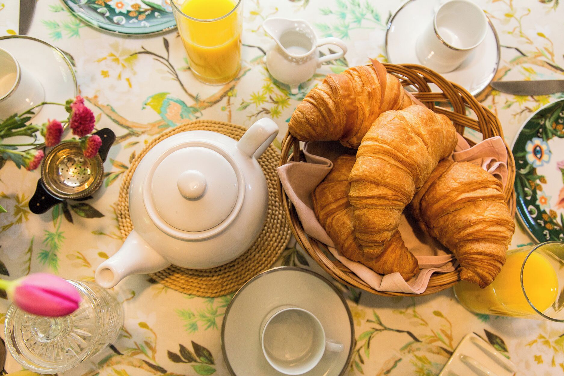 A breakfast setting with croissants, a teapot, cups, and a glass of orange juice.