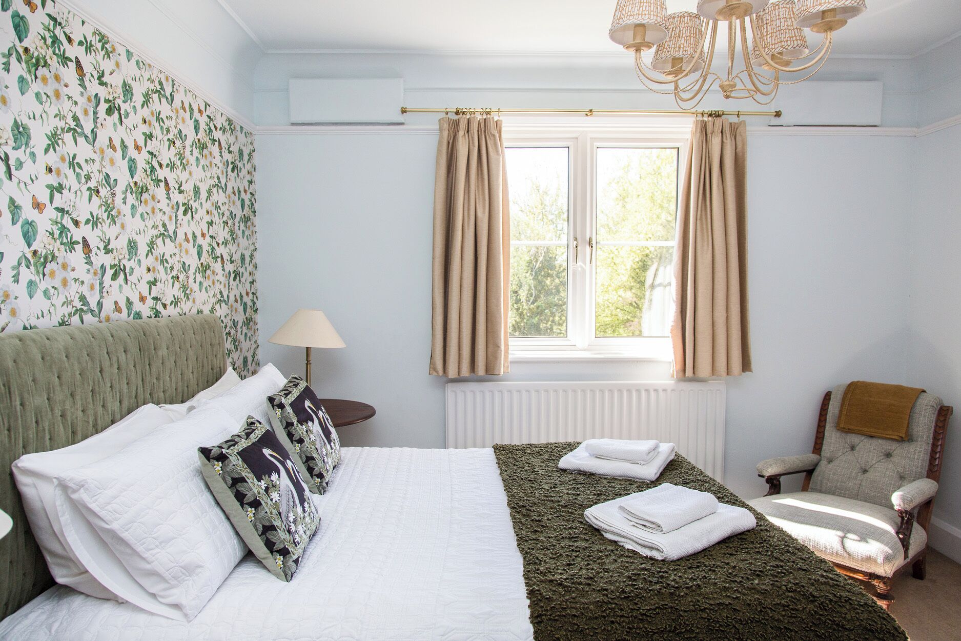 A bedroom with a bed, a chair, a window with curtains, a radiator, and a chandelier.